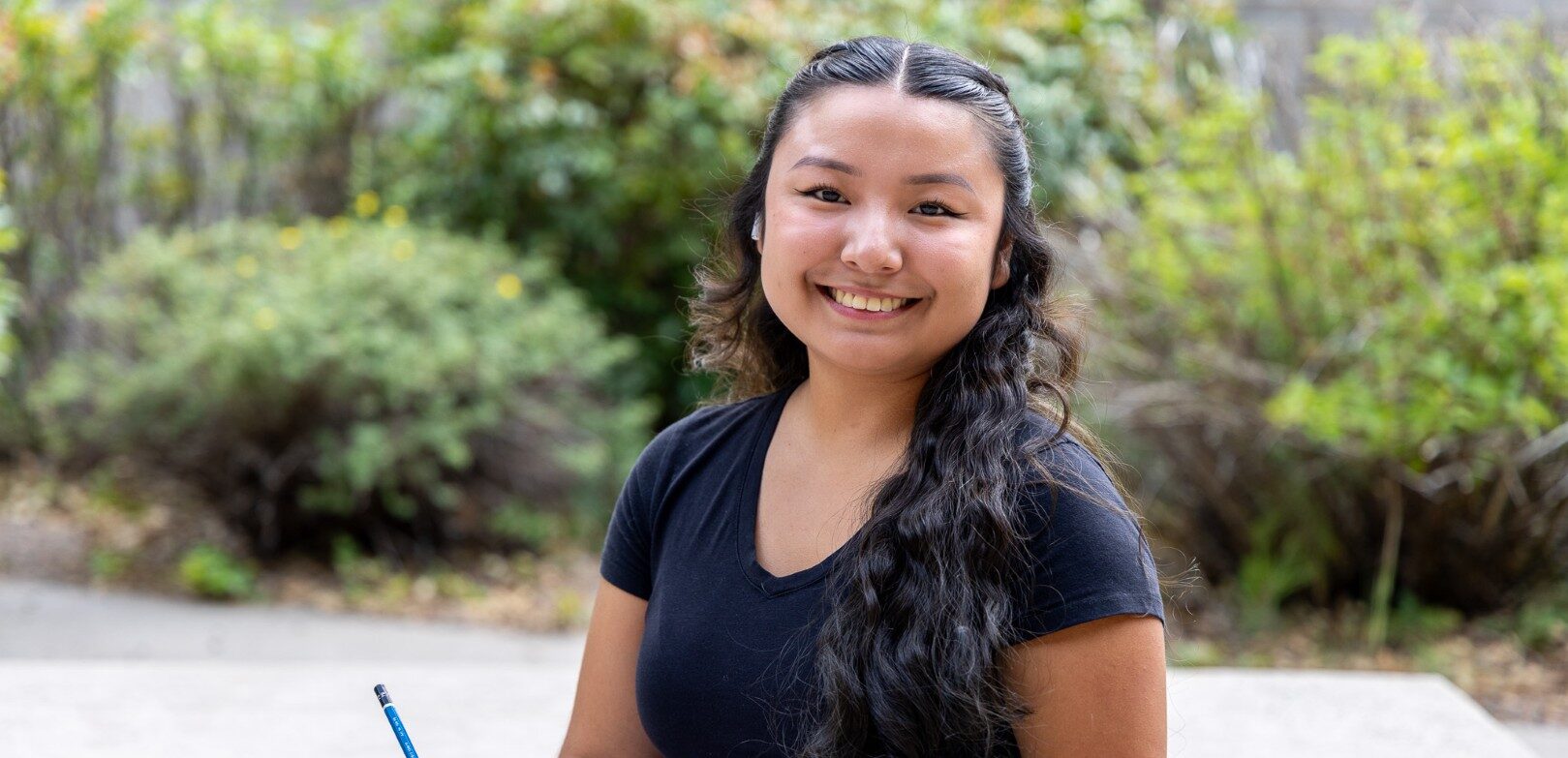 A smiling student sits outdoors on campus, holding an open notebook and pencil, with green shrubs in the background.