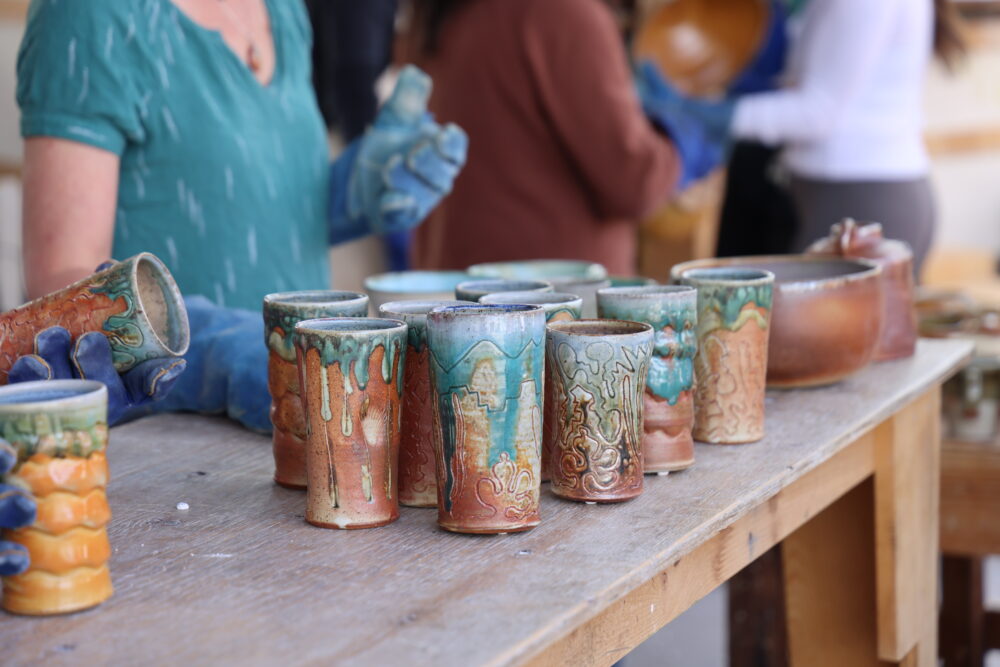 A collection of handmade ceramic cups with teal and rust glazes displayed on a wooden table during an outdoor pottery workshop, with participants visible in the background.