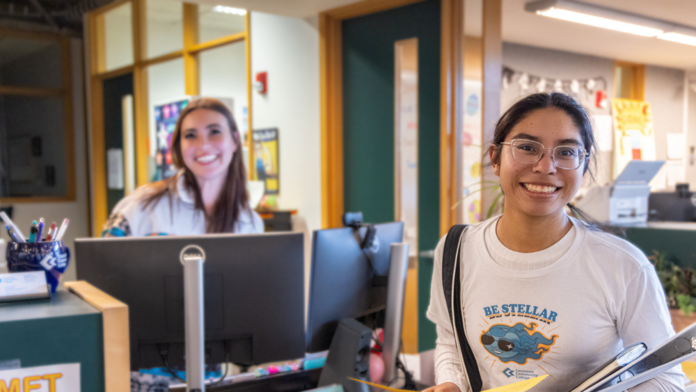 Student holding a binder stands at a campus service counter while connecting with staff in a student support office.