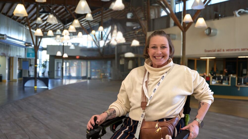 A CCC student using a power wheelchair smiles in the main atrium of the Lone Tree Campus, with the Financial Aid and Veterans Services office visible in the background.